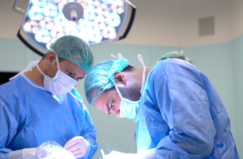 Two masked surgeons in blue gowns concentrating under OR light during a general-surgery procedure